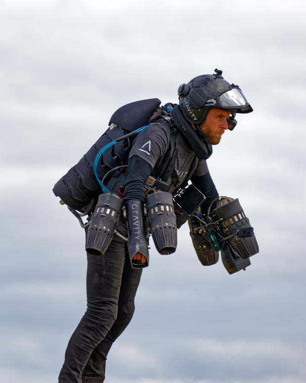 a man flies with the help of a very complicated looking jet pack.