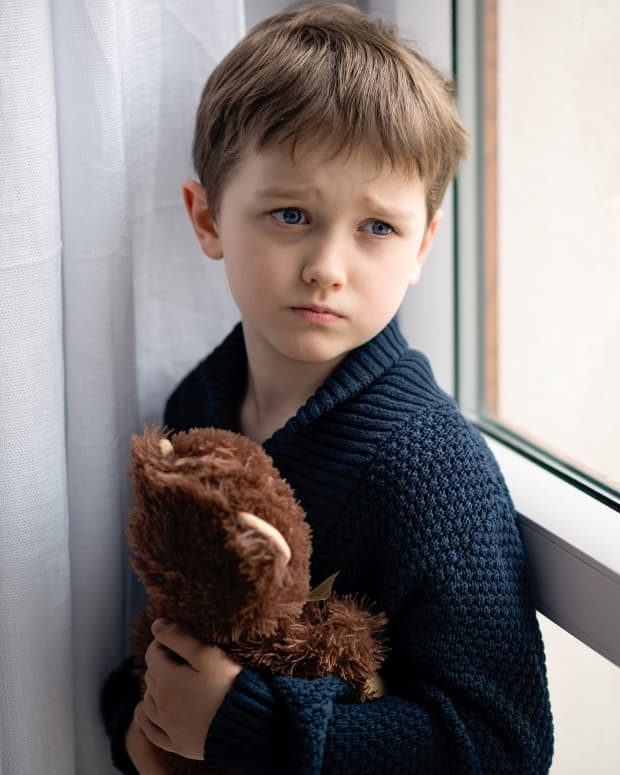 A boy huddles, frightened, with a teddy bear, behind a curtain near a window.