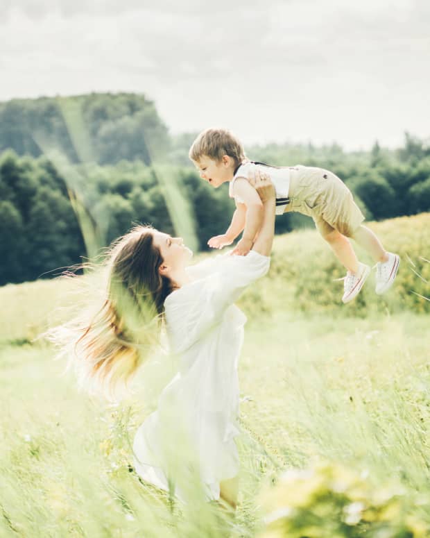 a white woman with long brown hair lifts a white boy child in the air in a grassy field while sunbeams shine down on them.